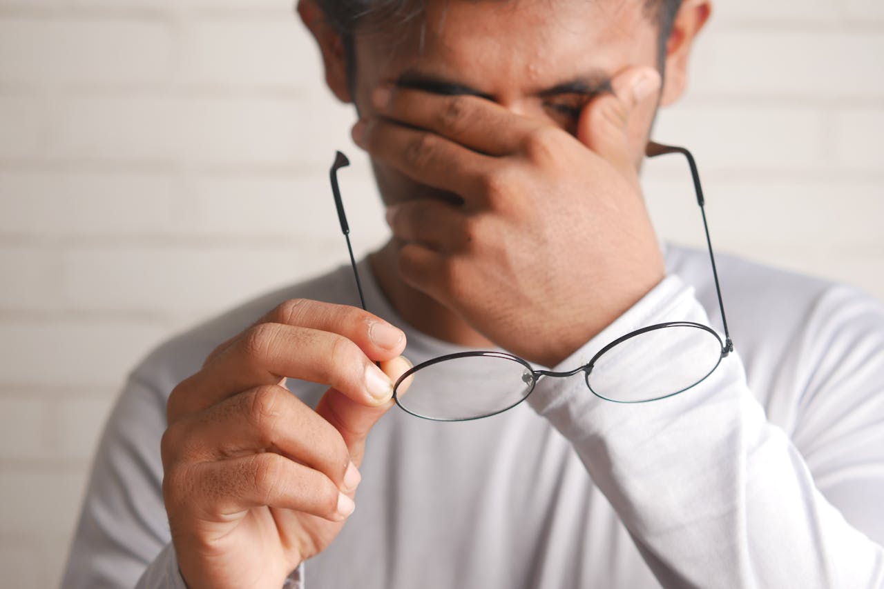 Close-up of a man holding eyeglasses, covering his face indoors, expressing stress or fatigue.